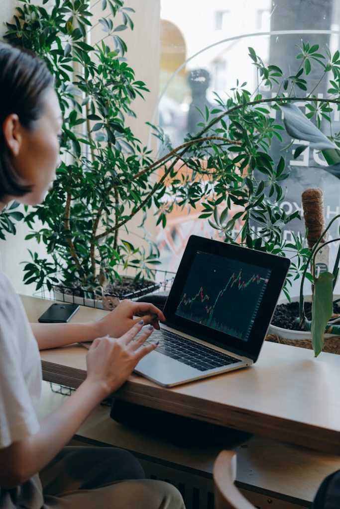 Businesswoman checks financial graphs on laptop in cozy café, surrounded by plants, showcasing remote work lifestyle.